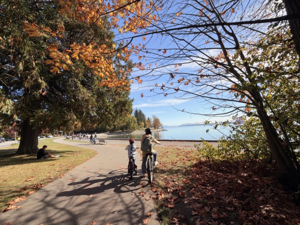 オレンジ色の紅葉と海を見ながら、スタンレーパークの道を自転車で進む親子