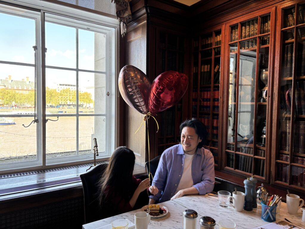 本棚に囲まれた部屋で朝ごはんを食べる父と娘。窓の外はテムズ川